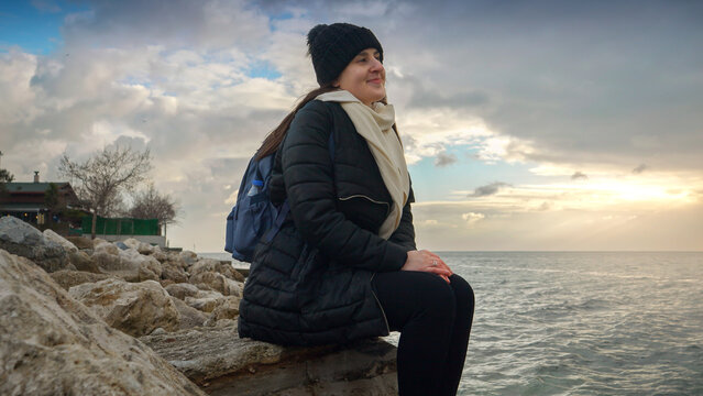 Happy smiling woman sitting on rocky beach and looking at cold sea waves - Powered by Adobe