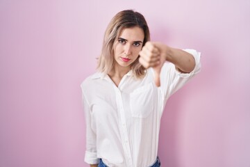 Young beautiful woman standing over pink background looking unhappy and angry showing rejection and negative with thumbs down gesture. bad expression.