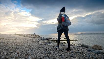 Fototapeta premium Young woman with a backpack and thermos takes a walk on a rocky sea beach, smiling as she explores the rugged terrain. Ideal for showcasing the journey and outdoor adventure.