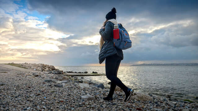 Female hiker in warm clothing equipped with a backpack and thermos, enjoying a walk on a rocky sea beach. A perfect addition to any travel or tourism project. - Powered by Adobe