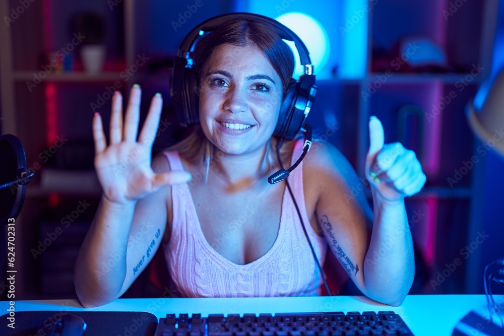 Poster Young blonde woman playing video games wearing headphones showing and pointing up with fingers number six while smiling confident and happy.