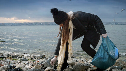 Slow motion of a woman in rubber gloves walking on the beach, picking up plastic waste and debris, and putting it in a garbage bag