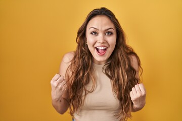 Young hispanic woman standing over yellow background celebrating surprised and amazed for success with arms raised and open eyes. winner concept.