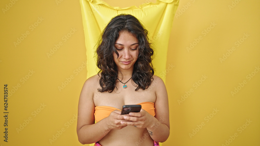 Wall mural Young beautiful hispanic woman tourist wearing bikini using smartphone over mattress float over isolated yellow background