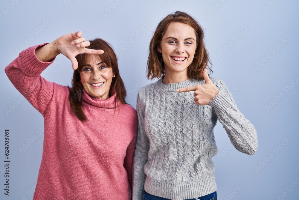 Poster Mother and daughter standing over blue background smiling making frame with hands and fingers with happy face. creativity and photography concept.