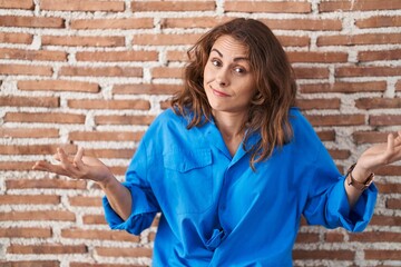 Beautiful brunette woman standing over bricks wall clueless and confused expression with arms and...