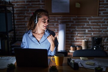 Beautiful brunette woman working at the office at night smiling with happy face looking and pointing to the side with thumb up.