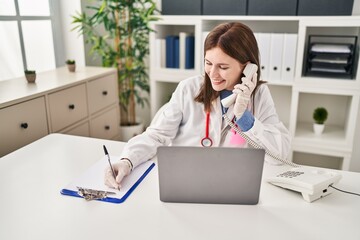 Young blonde woman doctor talking on telephone writing on document at clinic