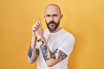 Young hispanic man with tattoos standing over yellow background holding symbolic gun with hand...