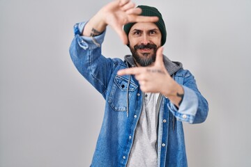 Young hispanic man with tattoos wearing wool cap smiling making frame with hands and fingers with happy face. creativity and photography concept.