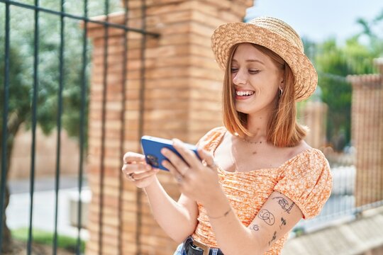 Young redhead woman tourist wearing summer hat watching video on smartphone at street