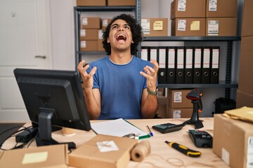 Hispanic man with curly hair working at small business ecommerce crazy and mad shouting and yelling with aggressive expression and arms raised. frustration concept.