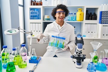 Hispanic man with curly hair working at scientist laboratory smiling happy pointing with hand and...