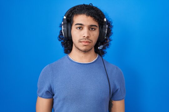 Hispanic man with curly hair listening to music using headphones relaxed with serious expression on face. simple and natural looking at the camera.