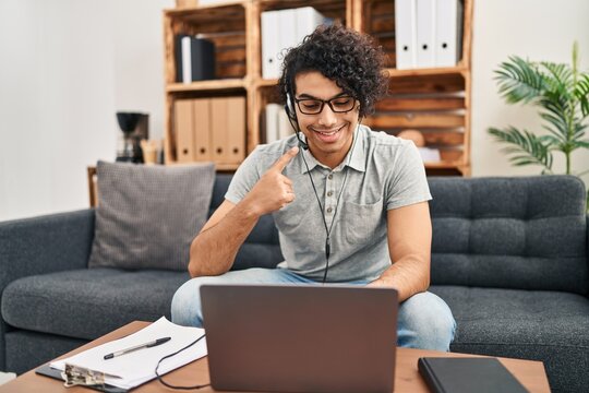Hispanic man with curly hair doing online session at consultation office smiling happy pointing with hand and finger