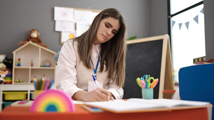 Young beautiful hispanic woman working as teacher writing at kindergarten