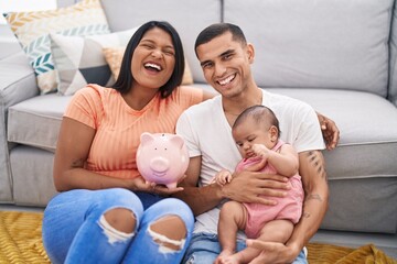 Young hispanic couple with baby holding piggy bank smiling and laughing hard out loud because funny...