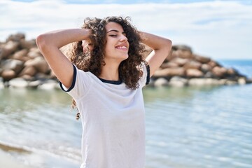 Young hispanic woman smiling confident relaxed with hands on head at seaside
