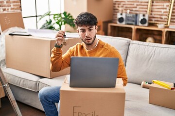 Hispanic man with beard holding keys of new home doing video call with laptop scared and amazed with open mouth for surprise, disbelief face