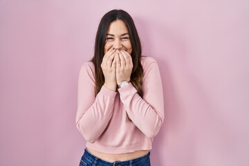 Young brunette woman standing over pink background laughing and embarrassed giggle covering mouth...