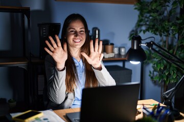 Young brunette woman working at the office at night showing and pointing up with fingers number nine while smiling confident and happy.