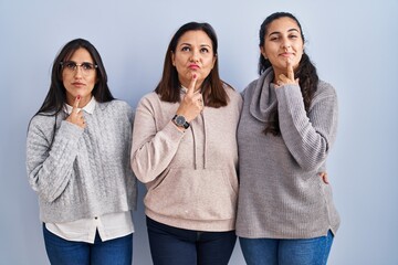 Mother and two daughters standing over blue background thinking concentrated about doubt with finger on chin and looking up wondering