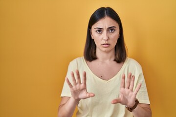 Obraz premium Hispanic girl wearing casual t shirt over yellow background moving away hands palms showing refusal and denial with afraid and disgusting expression. stop and forbidden.