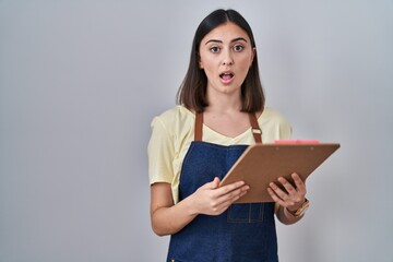 Hispanic girl wearing apron holding clipboard scared and amazed with open mouth for surprise, disbelief face