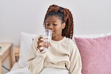 African american woman drinking glass of water sitting on bed at bedroom