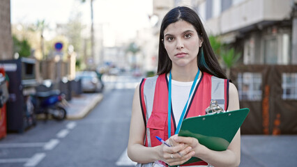 Young beautiful hispanic woman survey interviewer holding clipboard standing with serious expression at street