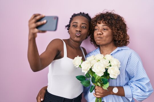 Two african women taking a selfie photo with flowers puffing cheeks with funny face. mouth inflated with air, catching air.