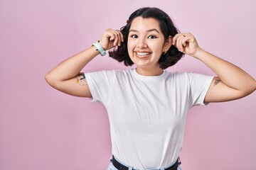 Young hispanic woman wearing casual white t shirt over pink background smiling pulling ears with fingers, funny gesture. audition problem