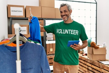 Middle age grey-haired man volunteer using touchpad holding clothes at charity center