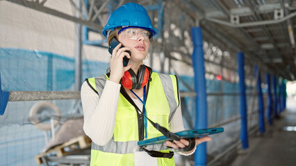 Young blonde woman architect talking on smartphone using touchpad at street