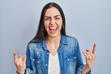 Hispanic woman standing over blue background shouting with crazy expression doing rock symbol with hands up. music star. heavy music concept.