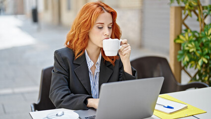 Young redhead woman business worker using laptop drinking coffee at coffee shop terrace