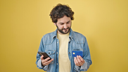 Young hispanic man shopping with smartphone and credit card over isolated yellow background