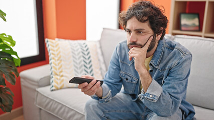 Young hispanic man watching tv sitting on sofa with boring expression at home