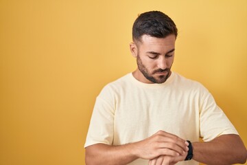 Handsome hispanic man standing over yellow background checking the time on wrist watch, relaxed and confident