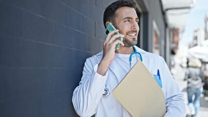 Young hispanic man doctor holding medical report talking on smartphone at hospital