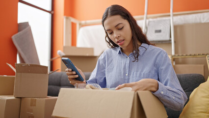 Fototapeta na wymiar Young beautiful hispanic woman unpacking cardboard box using smartphone at new home