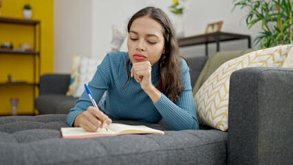 Young beautiful hispanic woman writing on notebook lying on sofa at home
