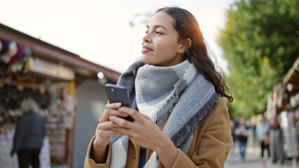 Fototapeta premium Young beautiful hispanic woman using smartphone smiling at street market