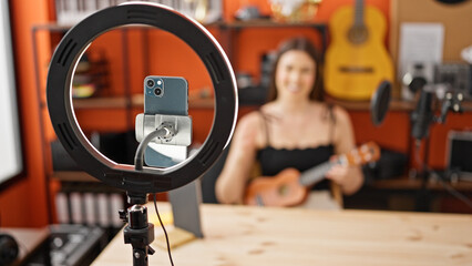 Young beautiful hispanic woman musician recording video tutorial playing ukulele at music studio