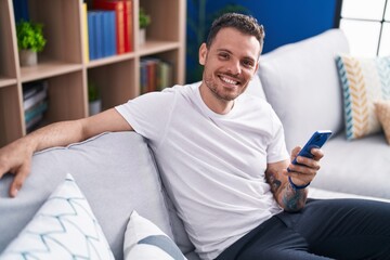 Young hispanic man using smartphone sitting on sofa at home