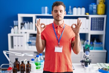 Young hispanic man working at scientist laboratory relax and smiling with eyes closed doing meditation gesture with fingers. yoga concept.