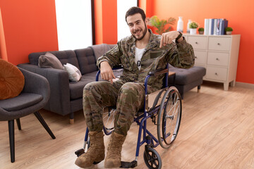 Young hispanic soldier man injured sitting on wheelchair smiling happy pointing with hand and finger