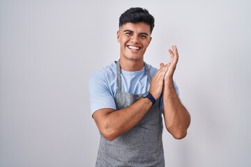 Hispanic young man wearing apron over white background clapping and applauding happy and joyful, smiling proud hands together