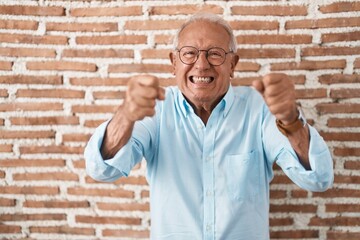 Senior man with grey hair standing over bricks wall angry and mad raising fists frustrated and...