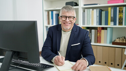 Middle age grey-haired man teacher using computer writing on notebook at university classroom